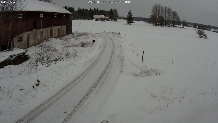 Landskap med snötäckt väg vid en gammal byggnad, omgiven av snöfält och träd i horisonten en vinterdag.