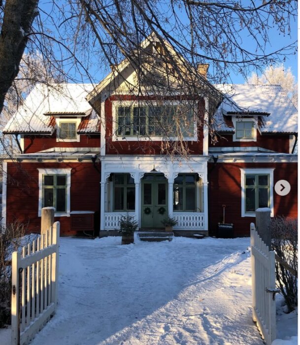 Rött trähus med vita detaljer och snötäckt gård, vintertid. Pittoresk entré och omgärdat av staket. Solen skiner genom tunna grenar.