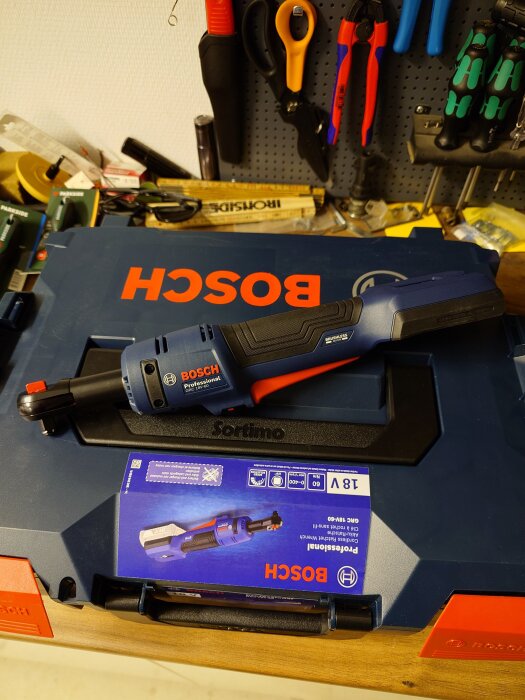 Blå Bosch batteridriven vinkelslip på en verktygslåda med verktyg som skruvmejslar och saxar hängande på en verktygstavla i bakgrunden.