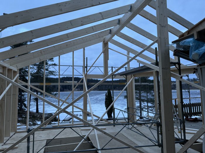 Träram för byggnation med takbalkar, delvis täckt av ställningar, med en sjö och skog i bakgrunden.