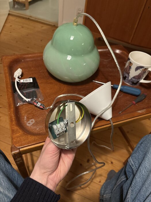 Hand som håller en metalltakkopp med sockerbit och jordad kabel; en grön lampa och verktyg syns i bakgrunden på en bricka.