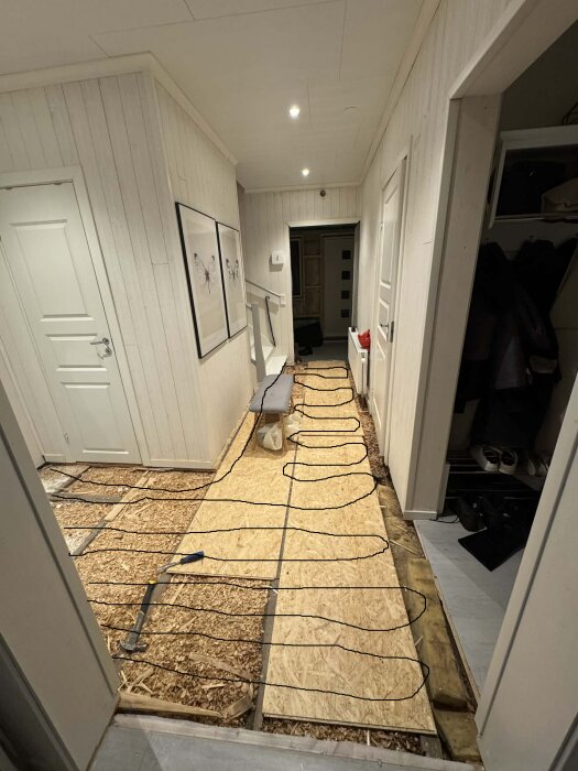 Entré med träskivor på golvet, del av golvprojekt, synliga spår för värmeslingor och en vägg med klädhängare och dörrar på sidorna.