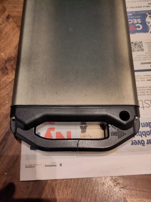 Sprucket handtag på ett lådcykelbatteri som ligger på ett bord med en tidning under.