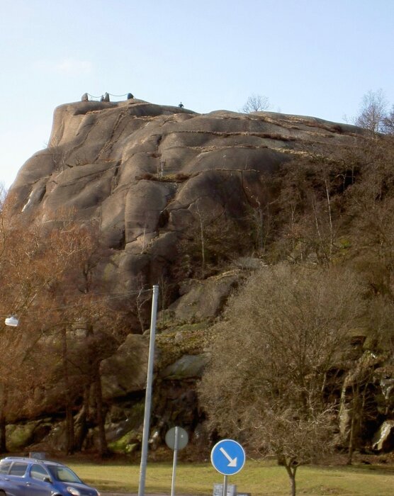 Bergsklippa med träd nedanför och människor på toppen, samt en vägskylt och bil i förgrunden.