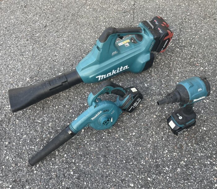 Tre blå Makita elverktyg med batterier på asfalt: en handdammsugare, en blåslampa och en mutterdragare.