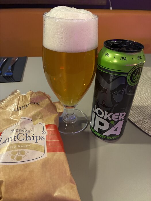 Ett glas öl med skum bredvid en Joker IPA-burk och en påse Svenska LantChips på ett bord.