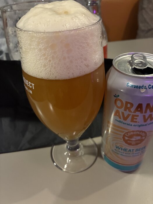 Ett glas fyllt med skummande veteöl bredvid en burk med texten "Orange Ave. Wit Wheat Beer".