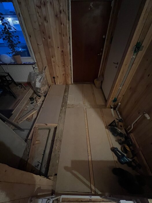 Byggarbetsplats i hall med brädor på golv, redskap vid sidan och en trädörr framför.