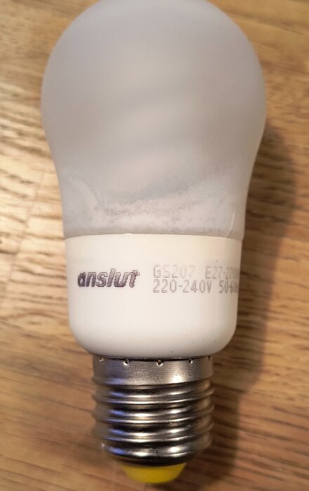 Lågenergilampa av märket Anslut liggande på ett trägolv, synlig text: "GS207 E27 220-240V 50-60Hz".