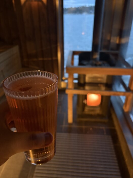 Hand håller i ett glas med Ankarudden Aussie Pale Ale i en vedbastu på Nynäs Havsbad, med utsikt över vattnet.