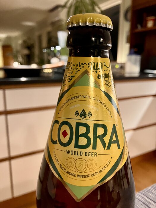 Flaska med Cobra World Beer, gul etikett med gröna detaljer, text "Global Multi-Award Winning Beer Medalist", i bakgrunden syns ett suddigt rum.