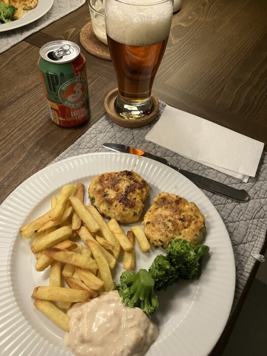 Tallrik med laxbiffar, pommes, broccoli och yoghurtblandning. Öl och läskburk vid sidan på bordet med underlägg och servett.
