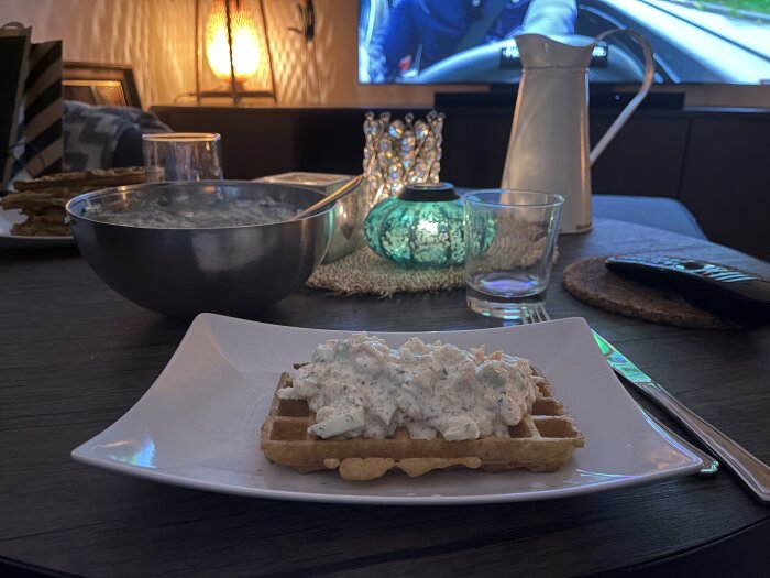 Potatisvåffla toppad med räkröra på en tallrik, omgiven av en skål med fyllning, ett glas och en karaff på ett bord.