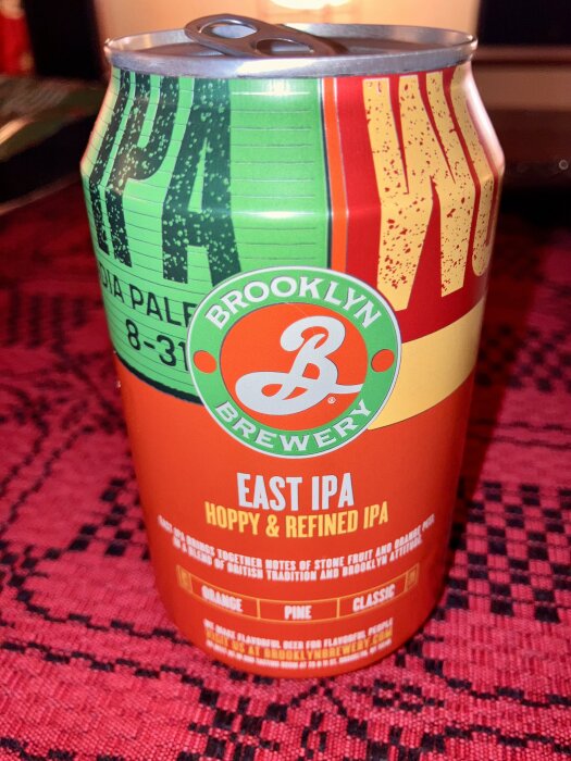 Burk med Brooklyn Brewery East IPA, står på röd duk. Färgglad design med text som beskriver smak och stil, inklusive citrus och tall.
