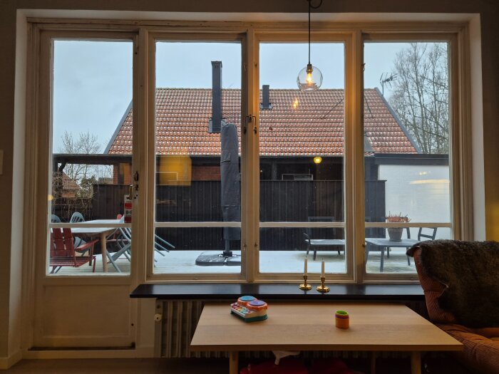 Stort fönsterparti med utsikt över en uteplats med bord och stolar, bakom syns en byggnad med tegelrött tak.
