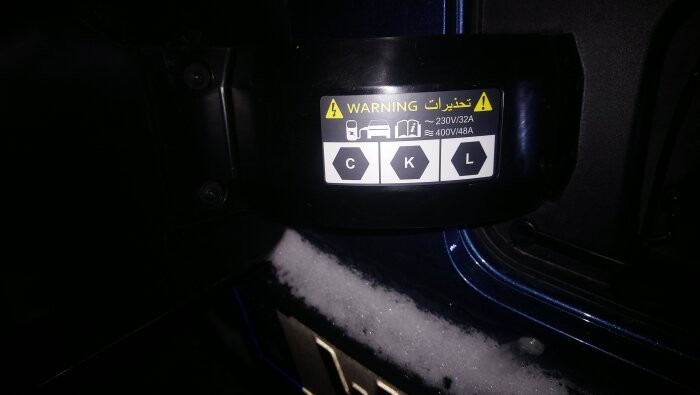 Varningsskylt med symboler och text på engelska och arabiska vid en laddport, omgiven av snö. Indikerar spänning och ampere.