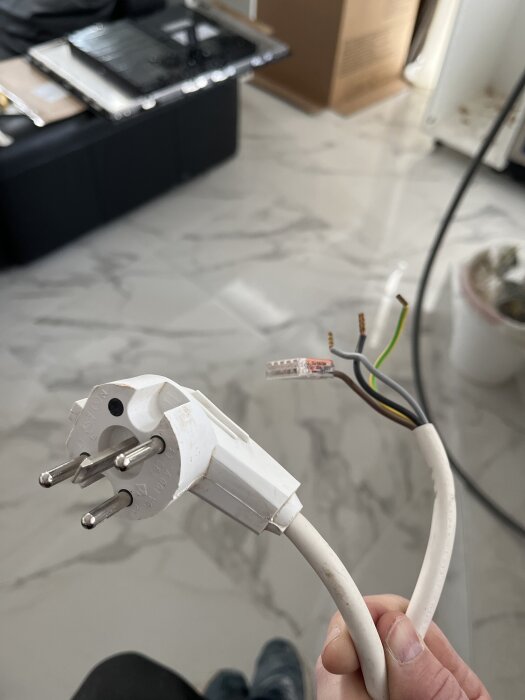 Vit stickkontakt med en trasig kabel, avslöjande tre ledningar, liggandes på ett marmorliknande golv.