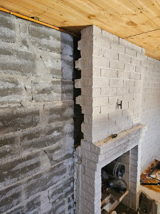 Renovering av öppen spis med vit tegelvägg intill grå cementvägg. Trätak ovanför, verktyg och eldsrör synliga i spisen.