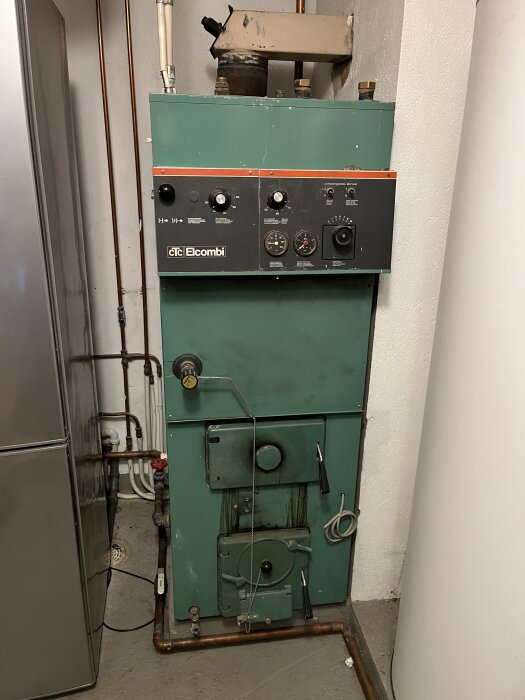 Äldre el-kombi-panna i grönt med panel och röranslutningar, står i ett pannrum med vit vägg och rörsystem.