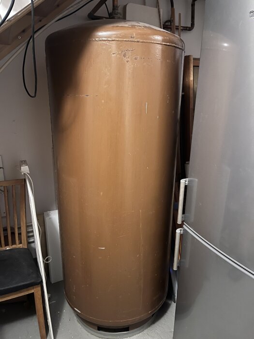 Brun, oisolerad tank i källare, placerad mellan en stol och ett kylskåp, med rör ovanför.
