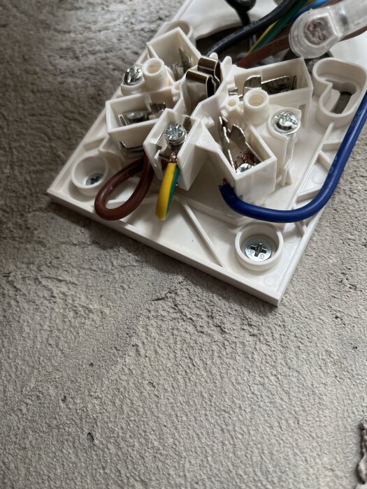 Elektrisk kopplingsdosa med brun, gul/grön och blå kabel ansluten på ett betonggolv.