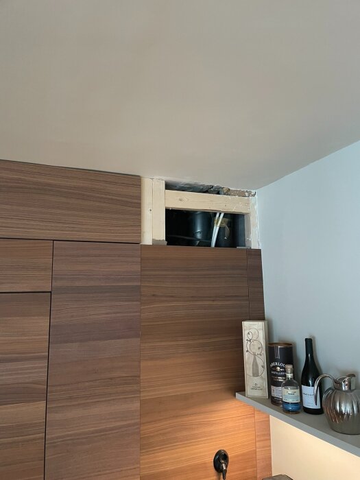 Öppning i köksskåp med synliga ventilationsrör och träreglar, täckt av bruna träpaneler, med dekorativa flaskor på en hylla intill.