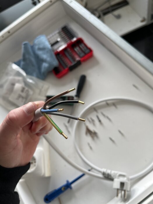 Hand som håller en bunt färgade elkablar, inklusive grå, framför en verktygslåda och andra verktyg på ett bord.