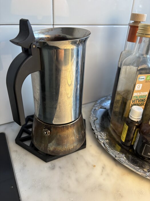 En espressobryggare i rostfritt stål står på en köksbänk bredvid flaskor med oljor och vinäger på ett silverfärgat fat.