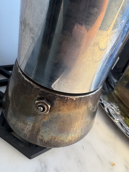 Närbild på en rostig och smutsig kaffepress med metalliskt utseende. Den står på en svart underlägg på en ljus bänkskiva.