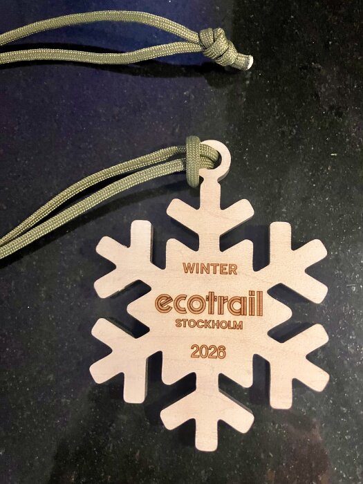 Träsnöflinga med text "Winter Ecotrail Stockholm 2026" och ett grönfärgat snöre, liggande på en mörk bakgrund.
