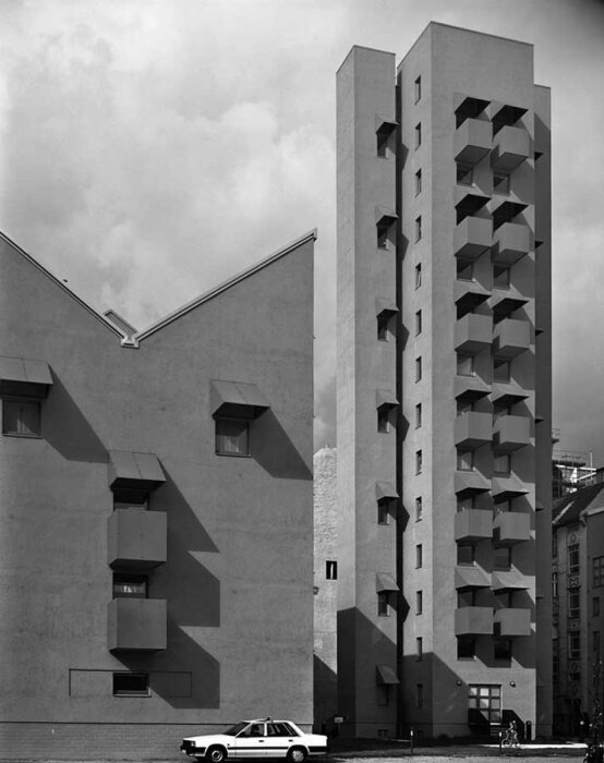 Svartvit bild av modernistiska byggnader med geometriska fönsteröppningar och en bil parkerad framför på en molnig dag.