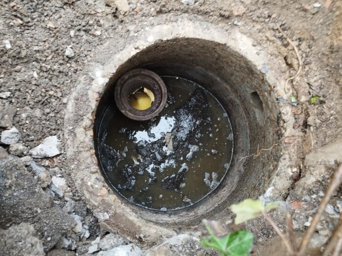 Öppen brunn med smutsigt vatten och en ansluten rördel, omgiven av jord och stenar.