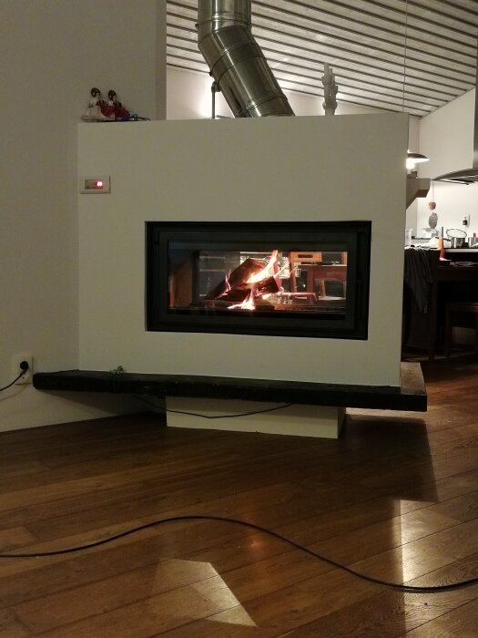 Eldstad med glaslucka och synlig eld, med metallrör för ventilation. Bakgrunden visar ett modernt kök med trädetaljer.