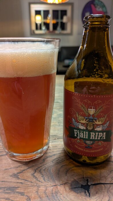 Ett glas fyllt med öl och en flaska märkt med Fjäll Ripa står på ett träbord. En suddig bakgrund syns i bakgrunden.