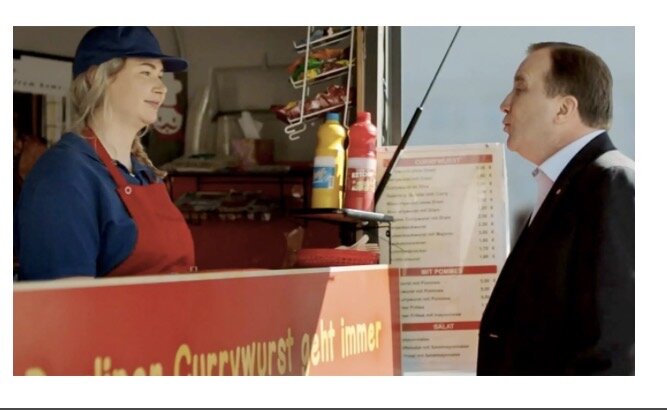 Två personer har en konversation vid en korvkiosk; menyn och korven syns i bakgrunden.