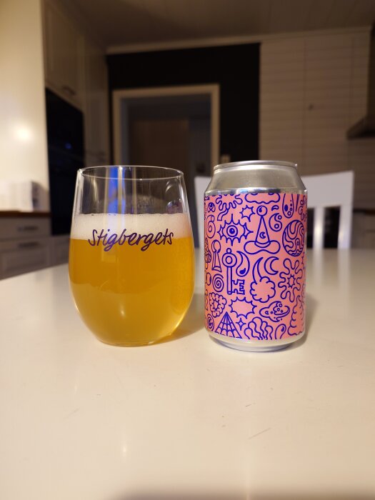 En ölburk med mönster bredvid ett glas med texten "Stigbergets", stående på ett bord i ett kök.