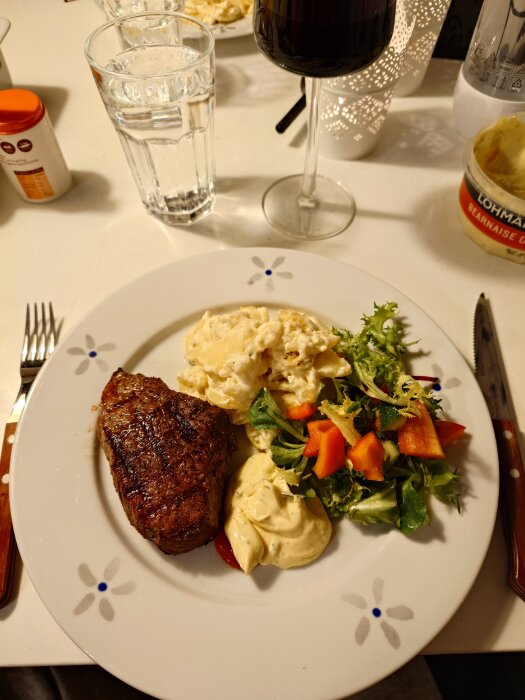 Tallrik med grillad köttbit, potatisgratäng och sallad med sås. Glas med rödvin och vatten bredvid samt bearnaisesås i burk.