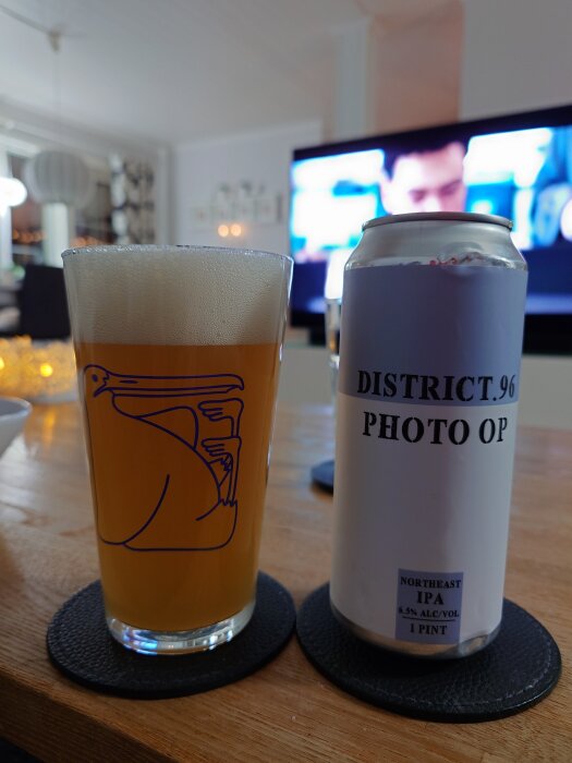 Glas öl med skum bredvid en ölburk med texten "District 96 Photo Op", på ett bord med suddig TV i bakgrunden.