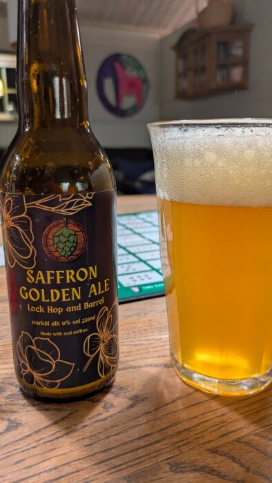 Flaska och glas med saffransöl på träbord, etikett med humlekotte och blommor, starköl 6% vol 330ml.