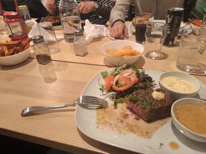 Tallrik med stek, sallad, såser och dryck på restaurangbord, omgiven av kryddor och fler serveringar i bakgrunden.