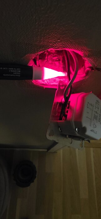 Elektrisk installation med en synlig dimmer, kablar och en ljuspenna i ett vägguttag, med ett rött sken på väggen och dimmern.