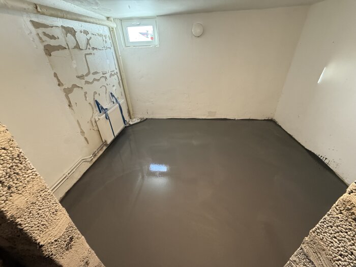 Rum med nygjuten betonggolv, omålade väggar och ett fönster. Värmeelement täckt med plast. Förberedelse för renovering.