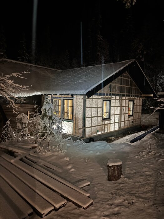 En byggnad under uppförande på en snötäckt tomt på natten, med synlig belysning och byggmaterial staplat på marken.