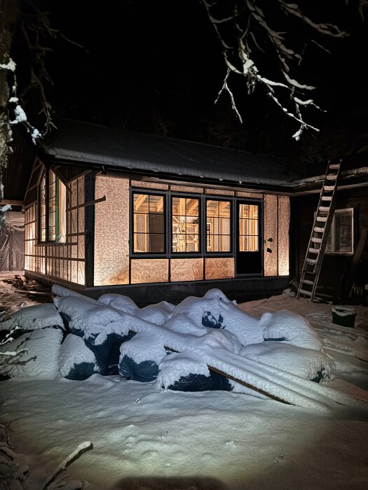 Snöigt bygge på natten med synliga takåsar och ångbroms. Omgivande snö täcker byggmaterial och en stege lutar mot huset.