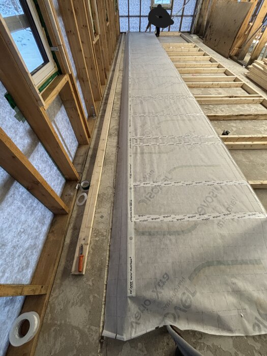 Installation av ångbroms på bygge där stomme av 45x95 reglar syns. Verktyg och tejp på golvet, fönsteröppningar med plywood synliga i bakgrunden.