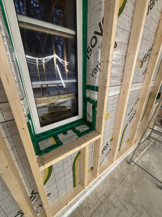 Väggkonstruktion med träreglar och fönster öppning, täckt med plastfolie och grönt tejp, synlig isolering och plywood runtom.
