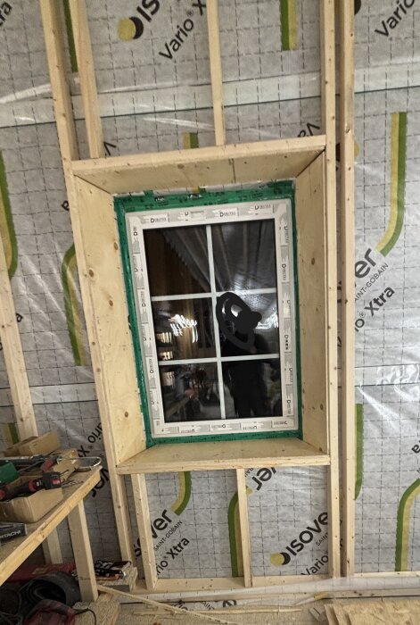 Fönsterinstallation i trävägg under konstruktion, med isoleringsmaterial synligt. Verktyg och arbetsmaterial ligger på en hylla bredvid.