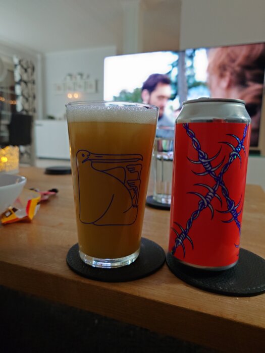 Glas med öl från Omnipollo på bord bredvid röd burk, en TV med oskarp scen syns i bakgrunden.