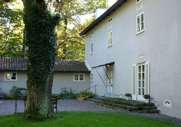 Exteriör av en vit villa med klassisk arkitektur, omgiven av träd och gräsmatta, ett exempel på 20-tals klassicism i Sverige.
