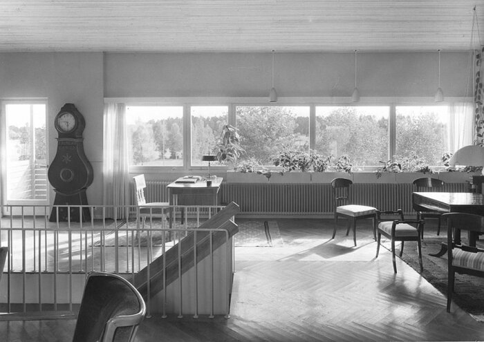 Ljust interiör av rum med fönstervägg, skrivbord, stolar och gammal klocka, utsikt mot trädgård. 20-talsklassicism.